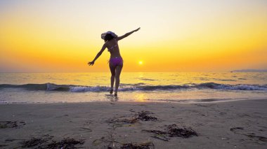 Boş bir plajda yürüyen, elleri açık ve özgürlüğün tadını çıkaran fit bir genç kız. Kız gün batımında deniz suyuna giriyor.
