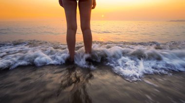 Sahilde gün batımında kadın ayaklarına su sıçrattı. Huzurlu ve rahatlatıcı okyanus suyu sahnesi güzel sahilde ayaklara sıçrıyor.