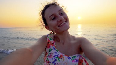 Bikinili güzel bir genç, arka planda yaz mevsiminin günbatımı ile komik selfie suratları yapar.