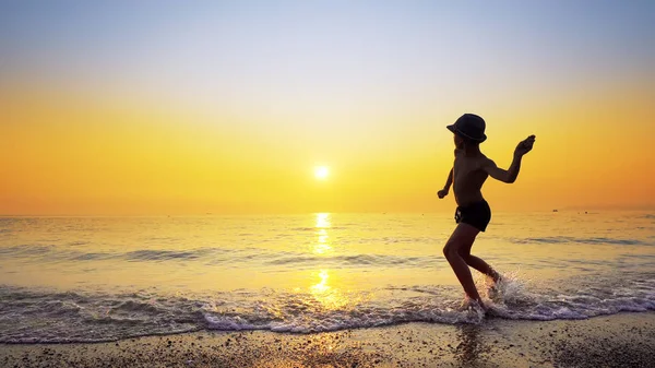 Şapkalı, taş atan, deniz suyu yüzeyinde zıplayan çocuk silueti. Canlı turuncu gökyüzü ile yaz tatili konsepti