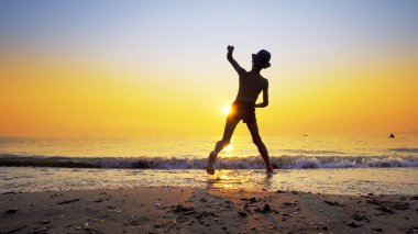 Şapkalı, taş atan, deniz suyu yüzeyinde zıplayan çocuk silueti. Canlı turuncu gökyüzü ile yaz tatili konsepti