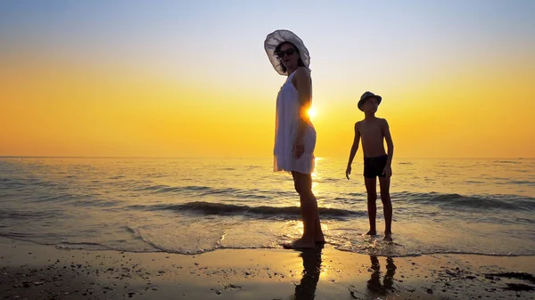 Happy family walk on sea coast and spraw water kicking the waves at sunset. Mother wear white dress and both have hats