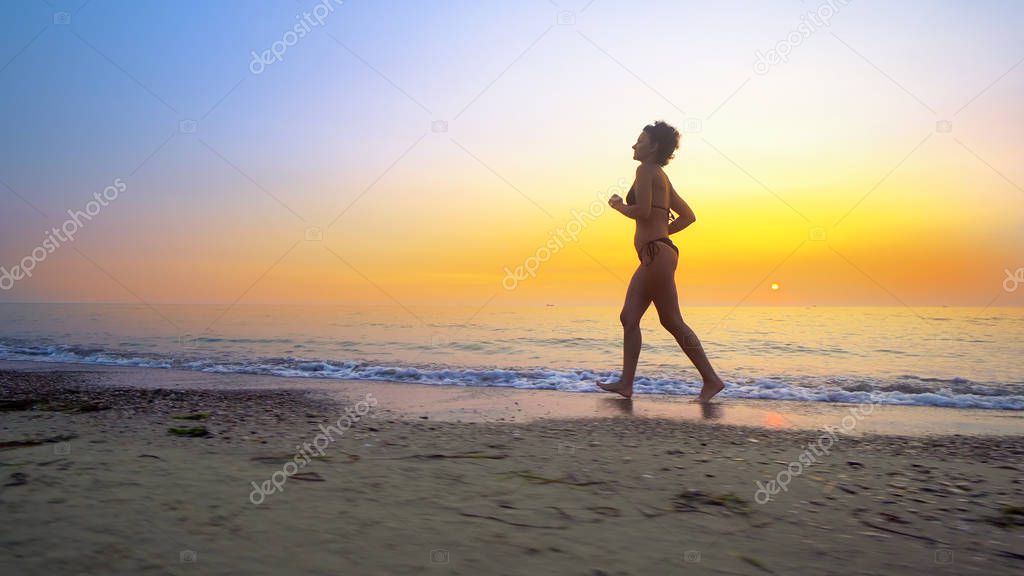 Barefoot sport woman in bikini jogging, running on an empty beach at sunset