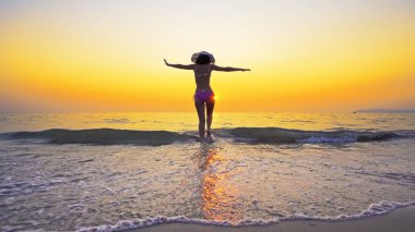 Bikinili ve şapkalı seksi genç gün batımında deniz suyundan çıkıyor, yaz tatili konsepti.