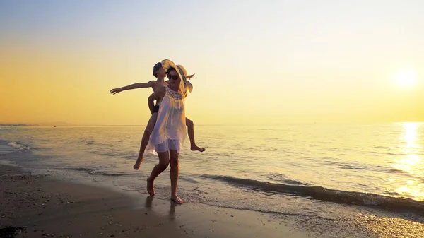 Mother gives her son a piggy back ride walking on empty ocean beach against sunset