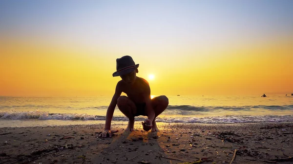 Silhouette of boy with hat throwing stones skipping on sea water surface. Summer vacation concept with vibrant orange sky