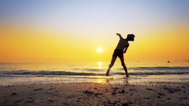 Şapkalı, taş atan, deniz suyu yüzeyinde zıplayan çocuk silueti. Canlı turuncu gökyüzü ile yaz tatili konsepti