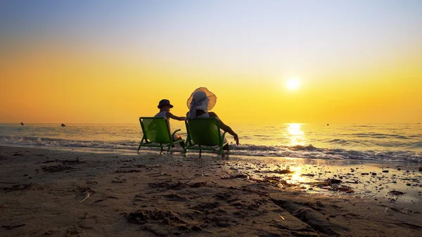 Happy family enjoy luxury sunset on the beach during summer vacations. Mother and son are sitting on a beach deck chair, against sunset, cinematic steadicam shot