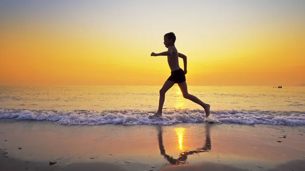 背景の夕日と海のビーチを走るスポーツ少年 — ストック写真