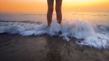 Sahilde gün batımında kadın ayaklarına su sıçrattı. Huzurlu ve rahatlatıcı okyanus suyu sahnesi güzel sahilde ayaklara sıçrıyor.