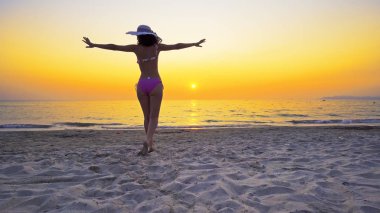 Boş bir plajda yürüyen, elleri açık ve özgürlüğün tadını çıkaran fit bir genç kız. Kız gün batımında deniz suyuna giriyor.