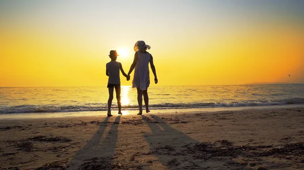 Family walk on beach to sea sunset and splash waves. Woman wear white dress and straw hat, boy has hat