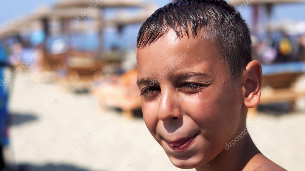 Handsome boy on beach look at camera and smile, the kid is dazzled by summer sun, cinematic dof