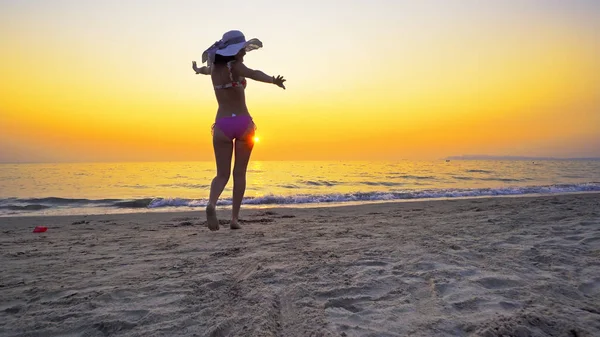 Fit female teen with hat walk on an empty beach with wide open hands enjoying freedom. Girl enter sea water at sunset