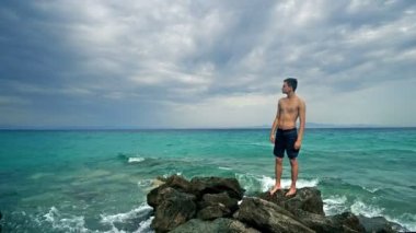 Fitness formundaki erkek sporcu deniz taşlarının üzerinde duruyor ve kaslarını gösteriyor.