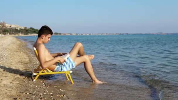 Νεαροί Άνδρες Διαβάζοντας Ένα Βιβλίο Στην Παραλία Κύματα Στο Παρασκήνιο — Αρχείο Βίντεο