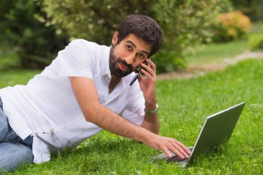 Genç mutlu gülümseyen adam dizüstü bilgisayar ve cep telefonu, açık havada çalışma