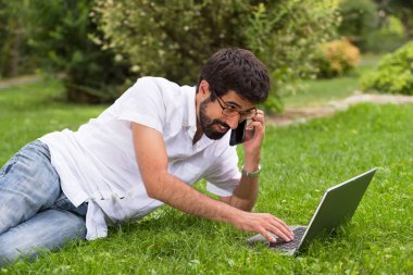 Genç mutlu gülümseyen adam dizüstü bilgisayar ve cep telefonu, açık havada çalışma