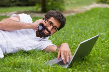 Genç mutlu gülümseyen adam dizüstü bilgisayar ve cep telefonu, açık havada çalışma