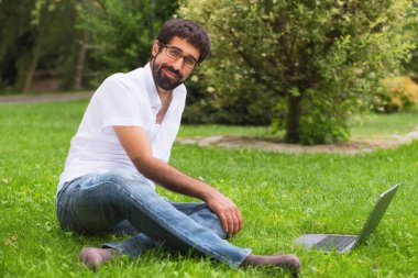 Parktaki genç adam çimlerin üzerinde dizüstü bilgisayarla oturuyor.