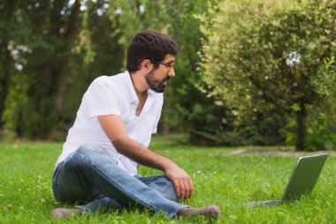Parktaki genç adam çimlerin üzerinde dizüstü bilgisayarla oturuyor.