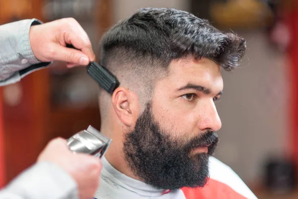 Красивый бородатый мужчина, стрижка парикмахером, электрический триммер в парикмахерской . — стоковое фото