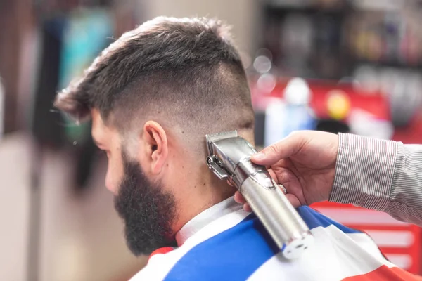 Handsome bearded man, getting haircut by barber, with electric trimmer at barbershop.