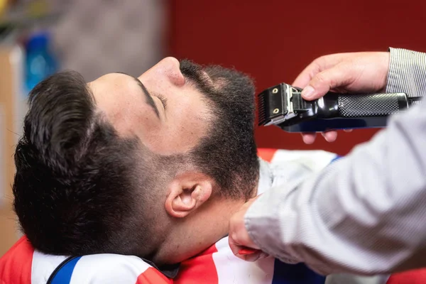 Kapper scheren van de baard van een knappe bebaarde man met een elektrisch scheerapparaat bij de kapper. — Stockfoto