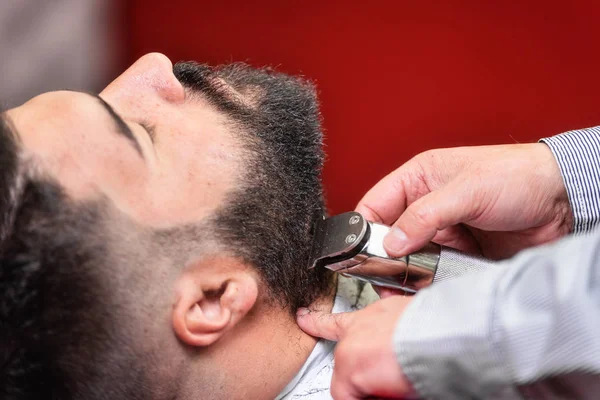 Kapper scheren van de baard van een knappe bebaarde man met een elektrisch scheerapparaat bij de kapper. — Stockfoto