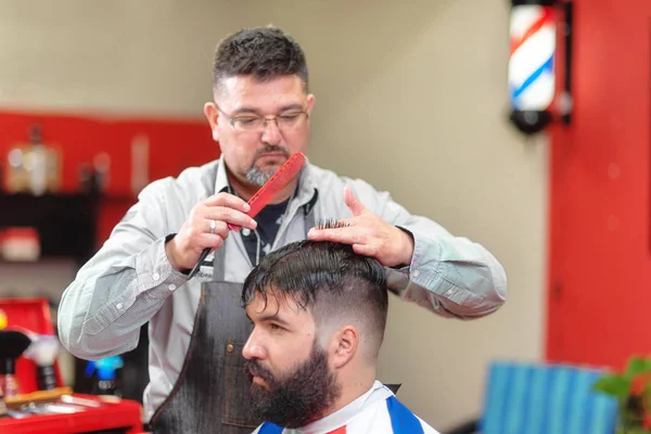 Bonito homem barbudo recebendo corte de cabelo por cabeleireiro na barbearia . — Fotografia de Stock