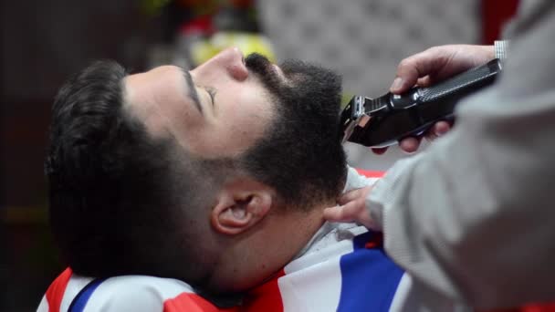 Peluquero afeitando la barba de un hombre barbudo guapo con una navaja eléctrica en la peluquería . — Vídeo de stock