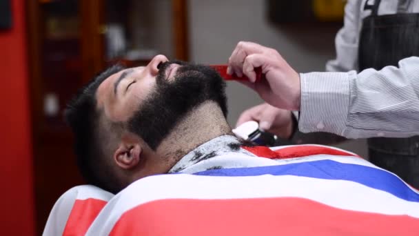 Peluquero afeitando la barba de un hombre barbudo guapo con una navaja eléctrica en la peluquería . — Vídeo de stock