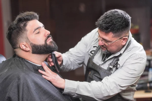 Kapper scheren van de baard van een knappe bebaarde man met een elektrisch scheerapparaat bij de kapper. — Stockfoto