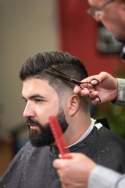 Handsome bearded man, having hair cut by scissors at barber shop.
