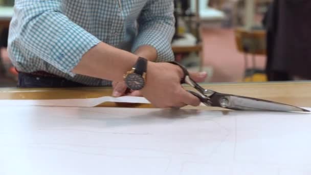 Tailor cutting out the marked pattern on fabric with large scissors on the workbench in his shop, close up view of his hands. — Stock Video