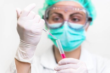 Koruyucu gözlüklü, maskeli ve eldivenli genç, çekici kadın bilim adamı laboratuardaki bir pipetle test tüpüne kırmızı sıvı madde atıyor..