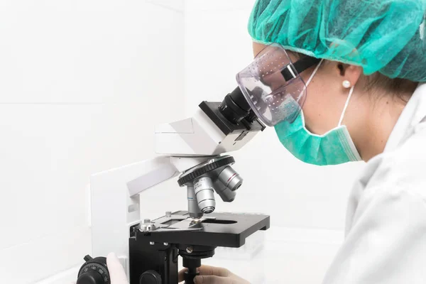 Jovem médica trabalhando com microscópio em laboratório, protegida contra riscos biológicos com máscara, chapéu e óculos . — Fotografia de Stock