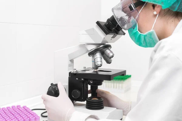 Młoda lekarka pracująca z mikroskopem w laboratorium, chroniona przed zagrożeniem biologicznym maską, kapeluszem i okularami. — Zdjęcie stockowe