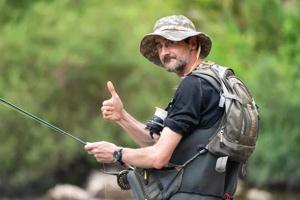釣り竿を手に川岸にあごひげを生やした幸せな漁師が親指を上げる。夏休み. — ストック写真