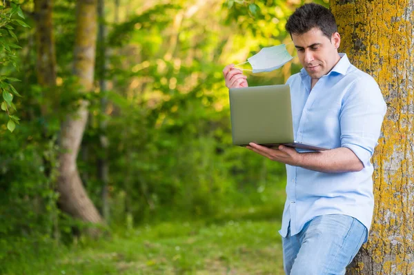 Handsome young man wearing face mask, standing in the park working using his laptop. — Stock Photo, Image