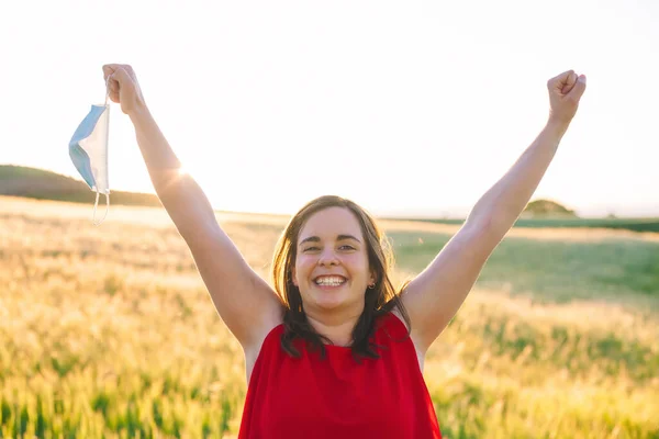 Happy smiling young woman, is standing with raised arms up, holding protective face mask in her hand. Woman enjoying breathing in nature outdoors. Celebrating end of Coronavirus pandemic. — Stock Photo, Image