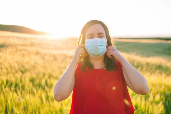 Szczęśliwa, wesoła młoda kobieta zdejmująca medyczną maskę na twarz, stojąca na łące podczas zachodu słońca wiosną. Koncepcja zakończenia pandemii koronawirusowej. — Zdjęcie stockowe