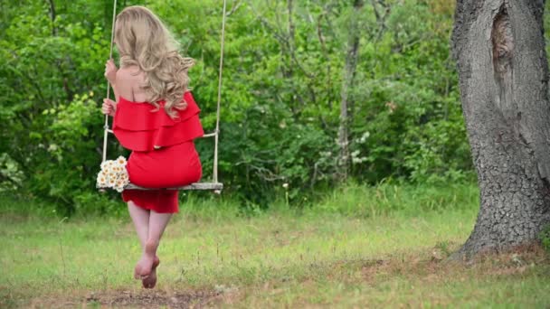 Egy piros ruhás nő hintázik a zöld nyári parkban. Csinos lány piros ruhában, hintázik. — Stock videók
