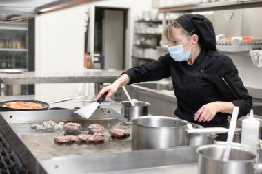 Koruyucu maskeli kadın şef bir restoranın ya da otelin mutfağında yemek hazırlıyor. Coronavirus önleme kavramı.