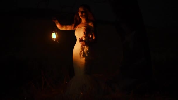 Femme effrayante avec une lanterne dans la scène de nuit Image effrayante d'une femme effrayante aux yeux sombres et l'apparence d'une sorcière, dans une robe blanche, tenant une lanterne allumée et une poupée effrayante. — Video