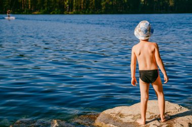 sevimli çocuk yüzme gövdeleri ve bir şapka beş yıllık bir güneşli yaz günü sahilde büyük bir kaya üzerinde duruyor. sırtını bir çocuktur
