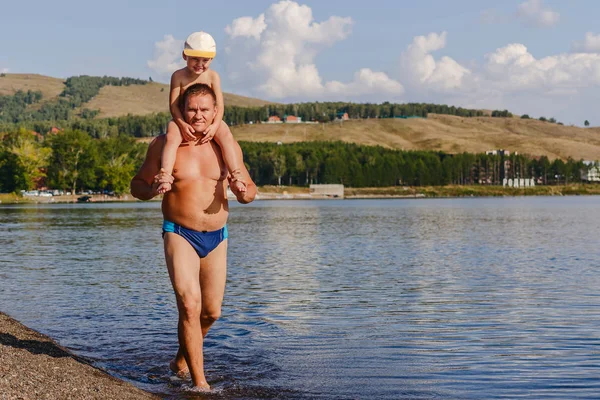 a man in swimming trunks carries a child on his neck on the lake in summer