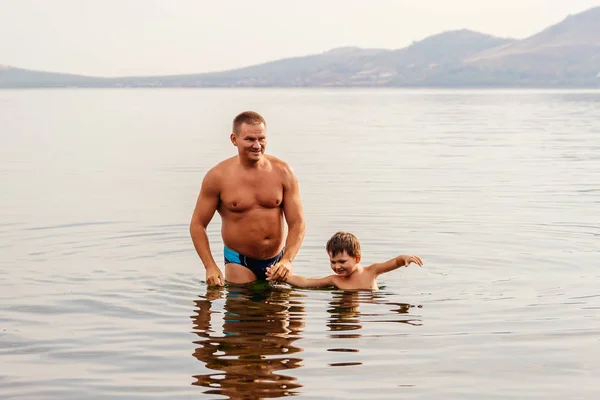 日焼けした父と水泳パンツ姿で幸せな息子は手を繋いでいる湖に立つ — ストック写真