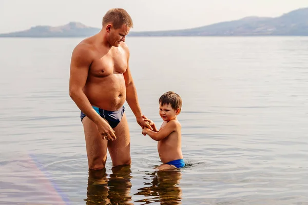 日焼けした父と息子の水泳パンツに手を繋いでいる湖に立つ — ストック写真