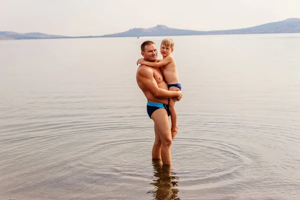 幸せな父は 湖に立っている間彼の幼い息子を抱擁します 父と息子の水泳パンツは — ストック写真